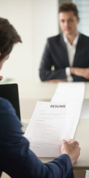 Back view of employer reading resume focus on document, job applicant waiting for hiring decision at desk on background. Hiring manager studying long list of candidates achievements and experiences